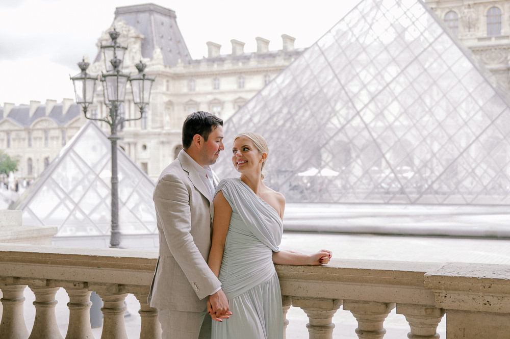 Couple leaning on a stone balustrade with the Louvre Pyramid and palace behind them in Paris